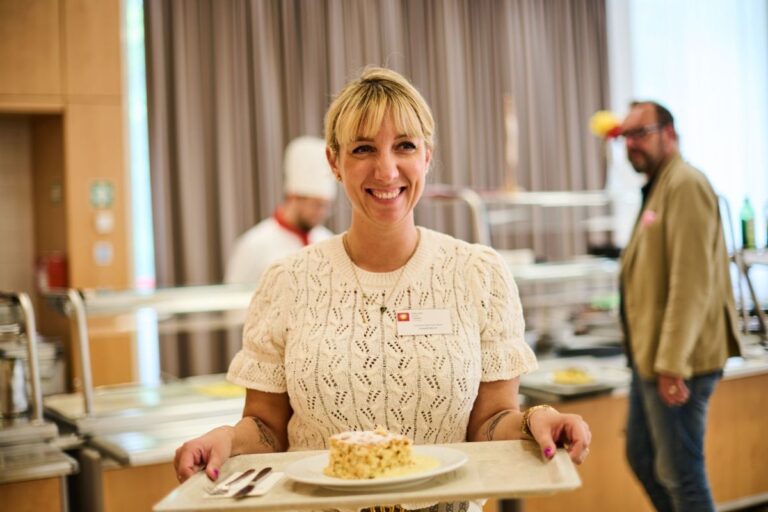 Frau mit Tablett und Dessert. Eine lächelnde Frau in weißem Strickpullover hält ein Tablett mit einem Dessert in der Hand, im Hintergrund eine Kantine mit Koch und Gästen.