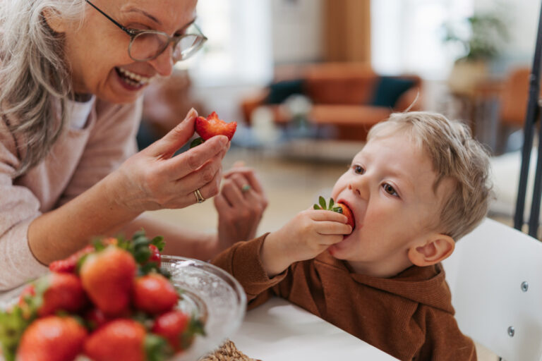 Eine ältere Frau hält lachend eine halbierte Erdbeere vor ein kleines Kind, das mit Genuss in eine andere Erdbeere beißt. Im Vordergrund steht eine Glasschale mit frischen Erdbeeren, im Hintergrund ein gemütlicher Wohnraum.