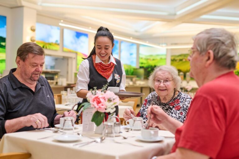 Serviererin bei Senior:innen: Eine junge Serviererin überreicht gut gelaunt ein Gericht an Senior:innen, die an einem festlich gedeckten Tisch sitzen.