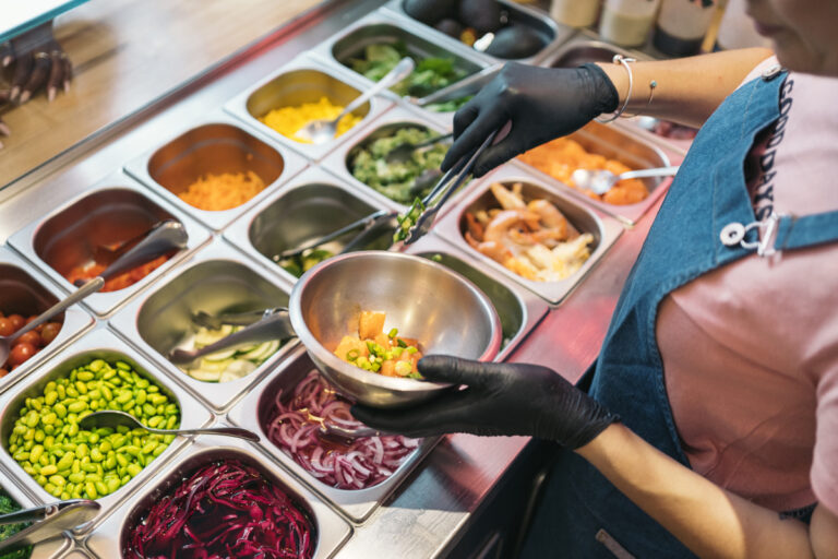 Eine Person mit blauen Handschuhen und Schürze füllt an einer Salatbar eine Bowl mit frischen Zutaten. In der Auslage befinden sich Edamame, rote Zwiebeln, Karotten, Gurken, Mais, Avocados, Shrimps und weitere geschnittene Gemüse.