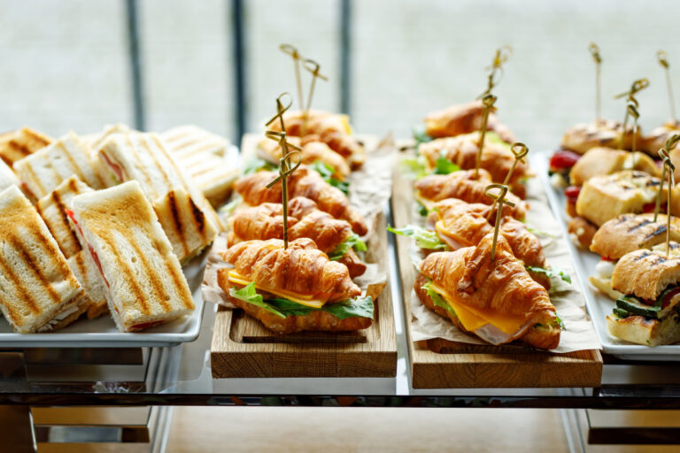 Buffet mit belegten Brötchen, Croissant-Sandwiches und Toast-Dreiecken, appetitlich angerichtet für Catering, Business-Lunch oder Event-Verpflegung. Frische Zutaten wie Käse, Schinken, Salat und Grillgemüse auf dekorativen Holzplatten serviert.