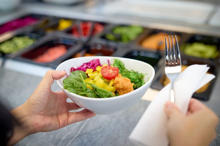 Eine Hand hält eine Schüssel mit verschiedenen Salaten, im Hintergrund ein Buffet.